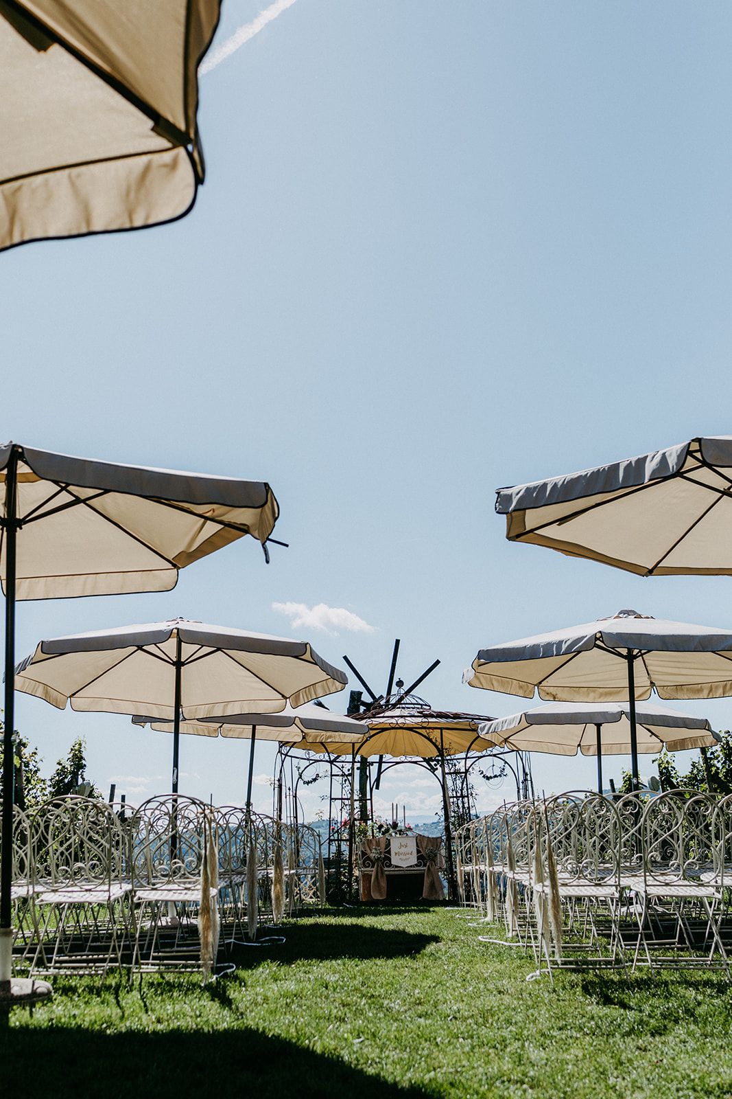 Outdoor wedding ceremony setup with white umbrellas and chairs on green lawn under blue sky.