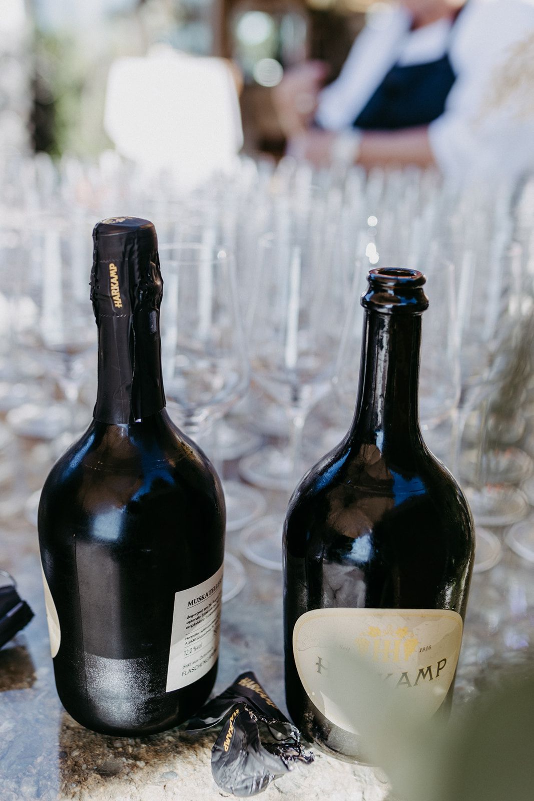 Two dark wine bottles displayed beside empty champagne glasses at outdoor wedding reception.