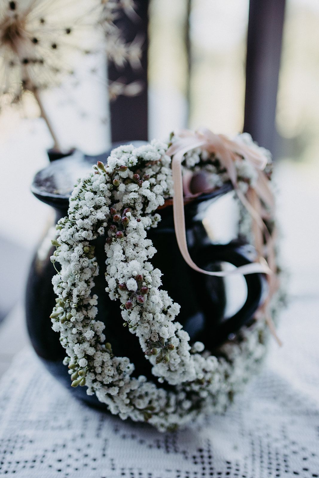Delicate white baby's breath flower crown with pink ribbon resting on a dark ceramic vase.