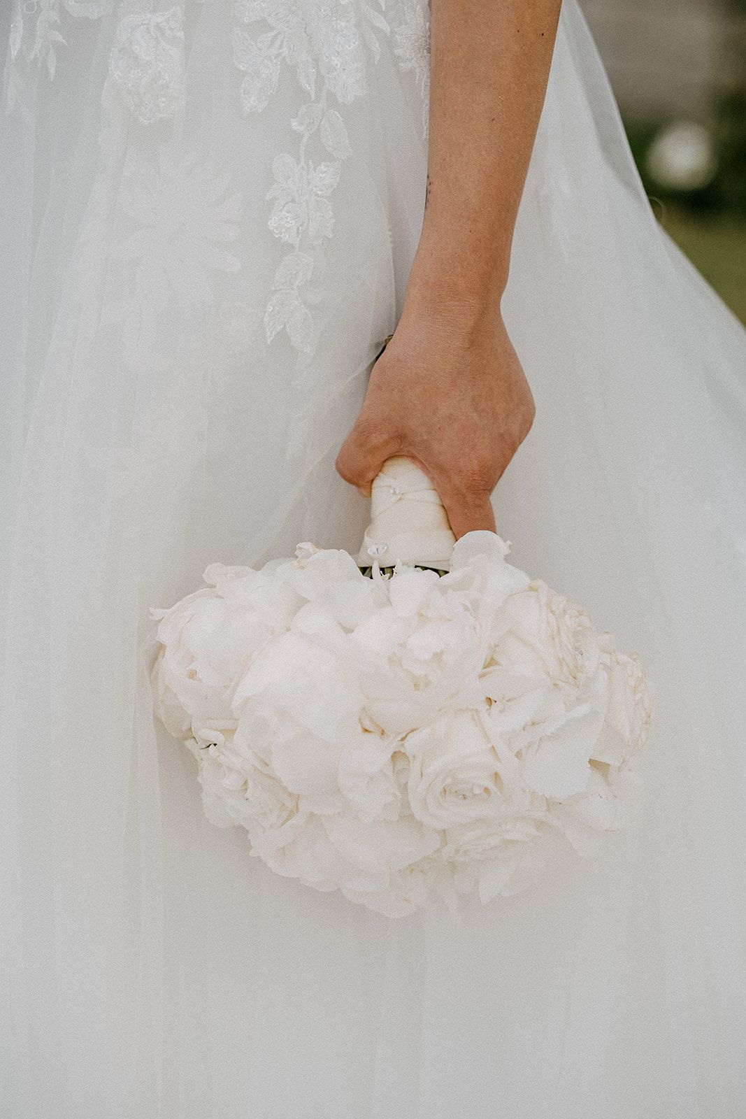 Close-up of bride holding elegant white peony and rose wedding bouquet against lace gown.