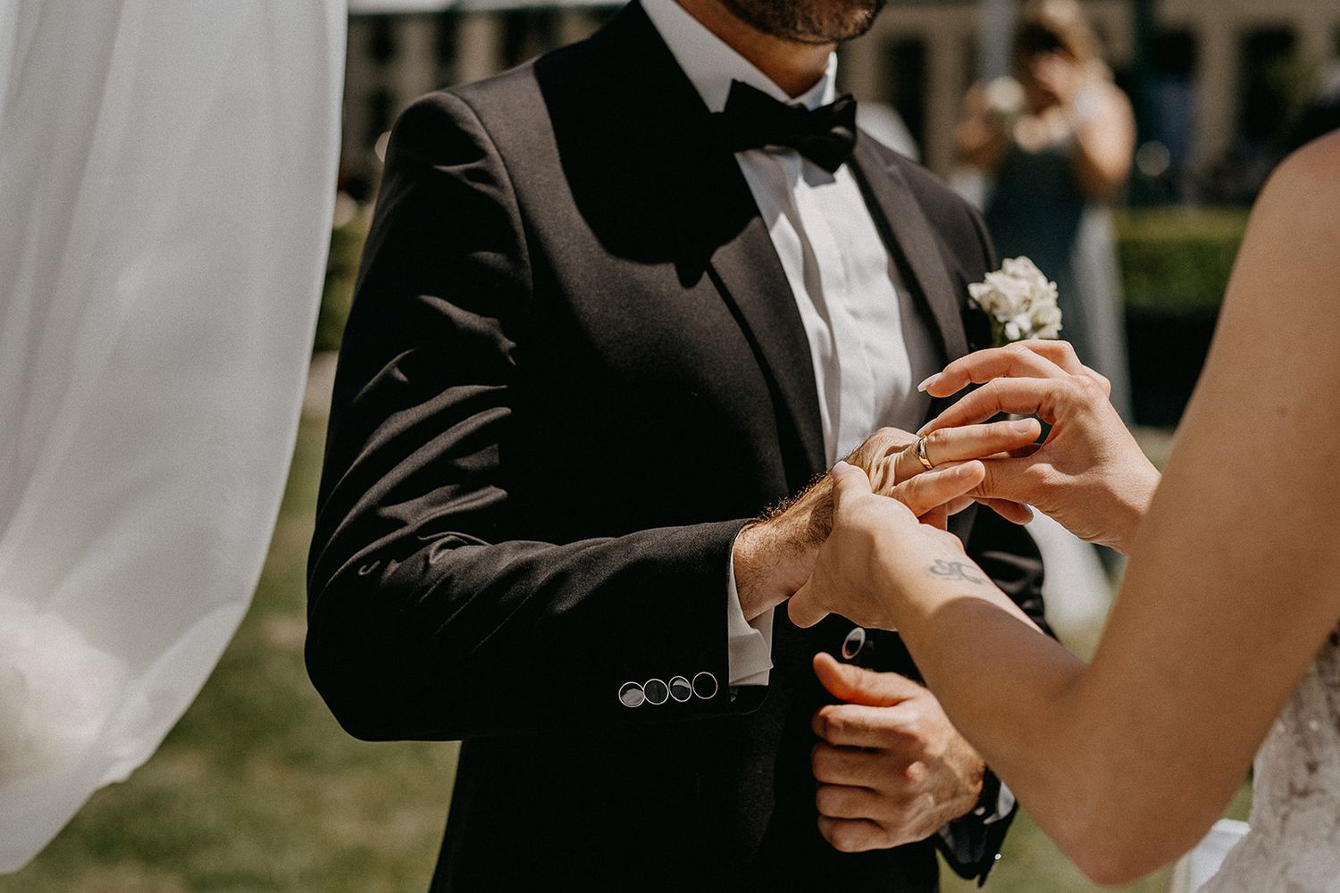 Groom in black tuxedo exchanges wedding rings with bride during elegant outdoor wedding ceremony.