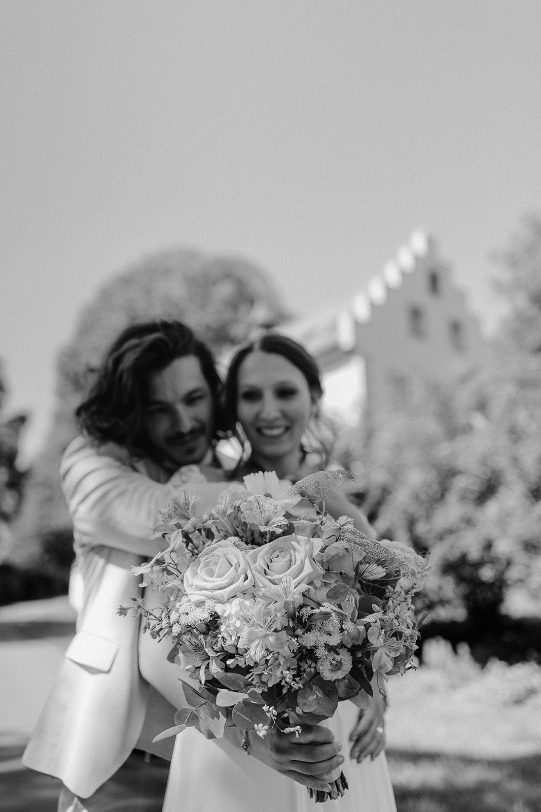Black and white photo of groom embracing bride from behind as she holds a floral bouquet.