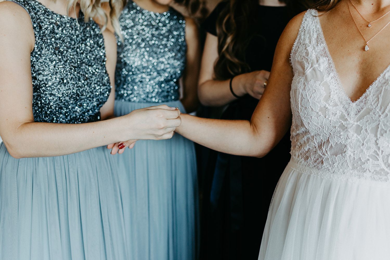 Close-up of bridesmaids in blue sequin dresses holding hands with bride in lace gown.
