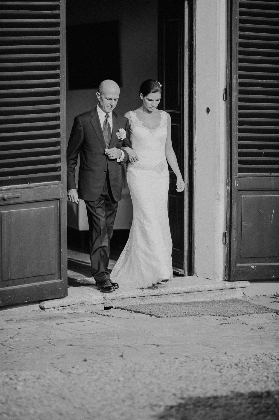 Black and white photo of bride in fitted gown escorted by older man through wooden doors. Tuscany Villa Catignano