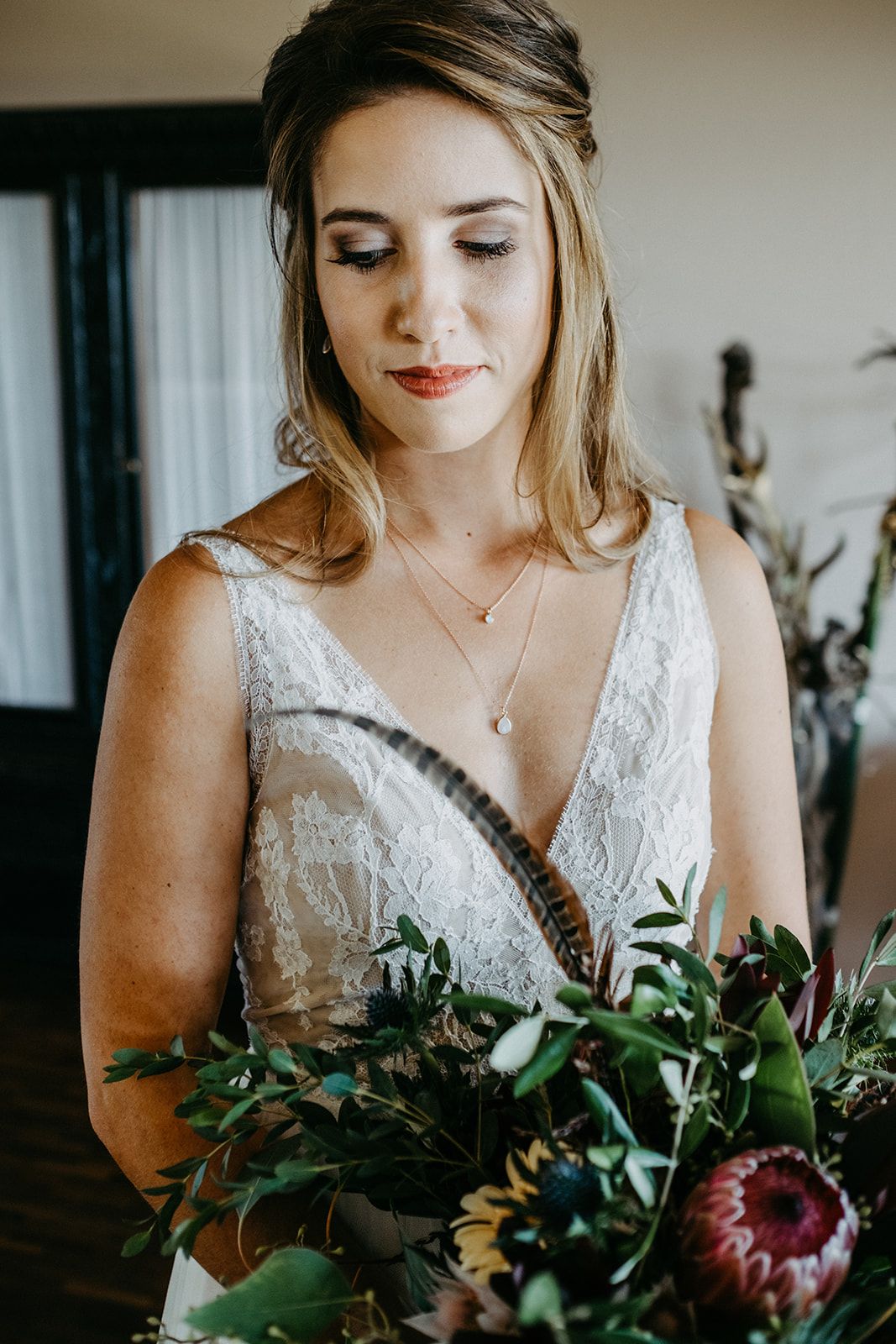 Elegant bride in lace V-neck wedding dress holding lush floral bouquet with protea and greenery.