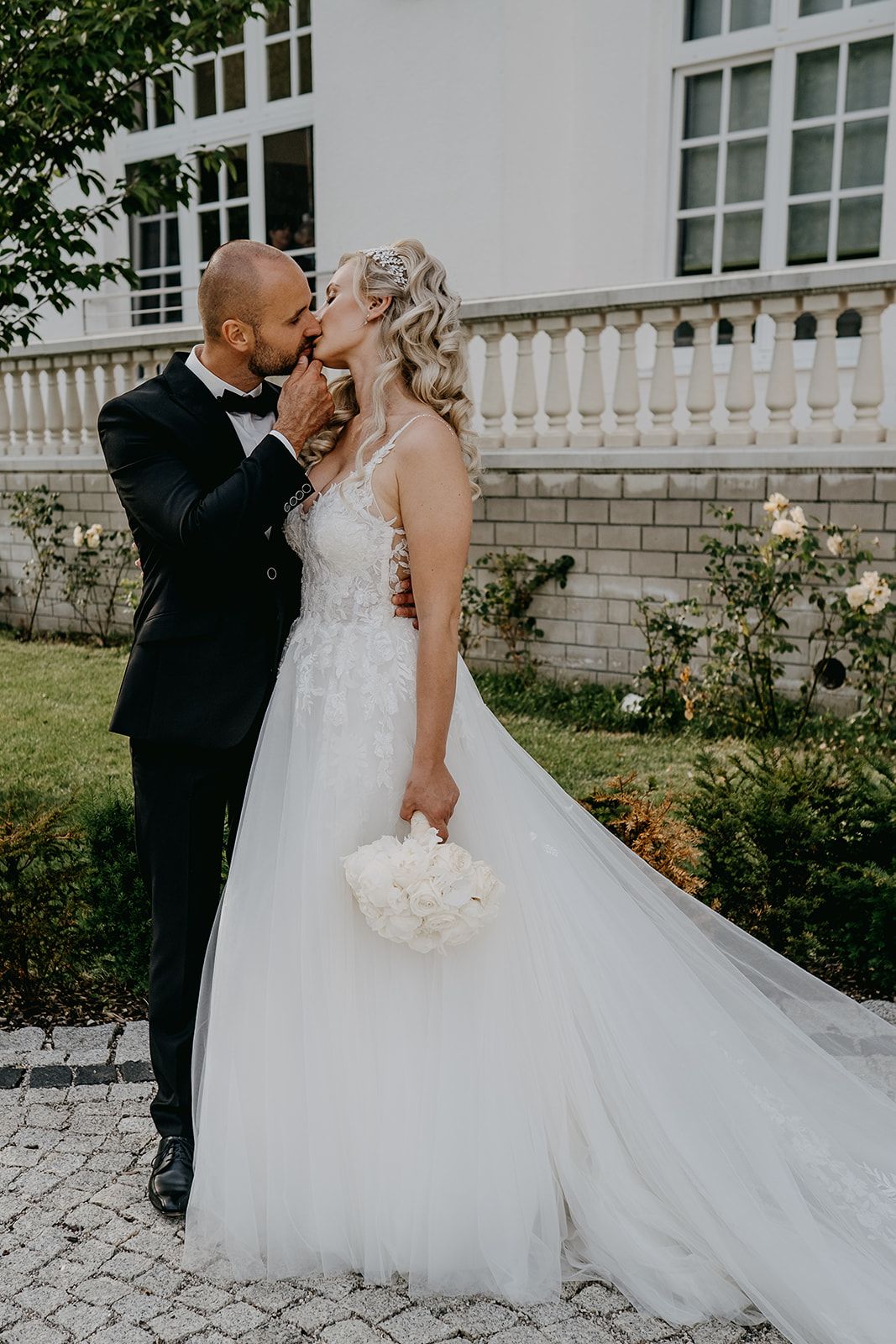 Bride and groom share romantic kiss outdoors at elegant white manor house wedding venue.