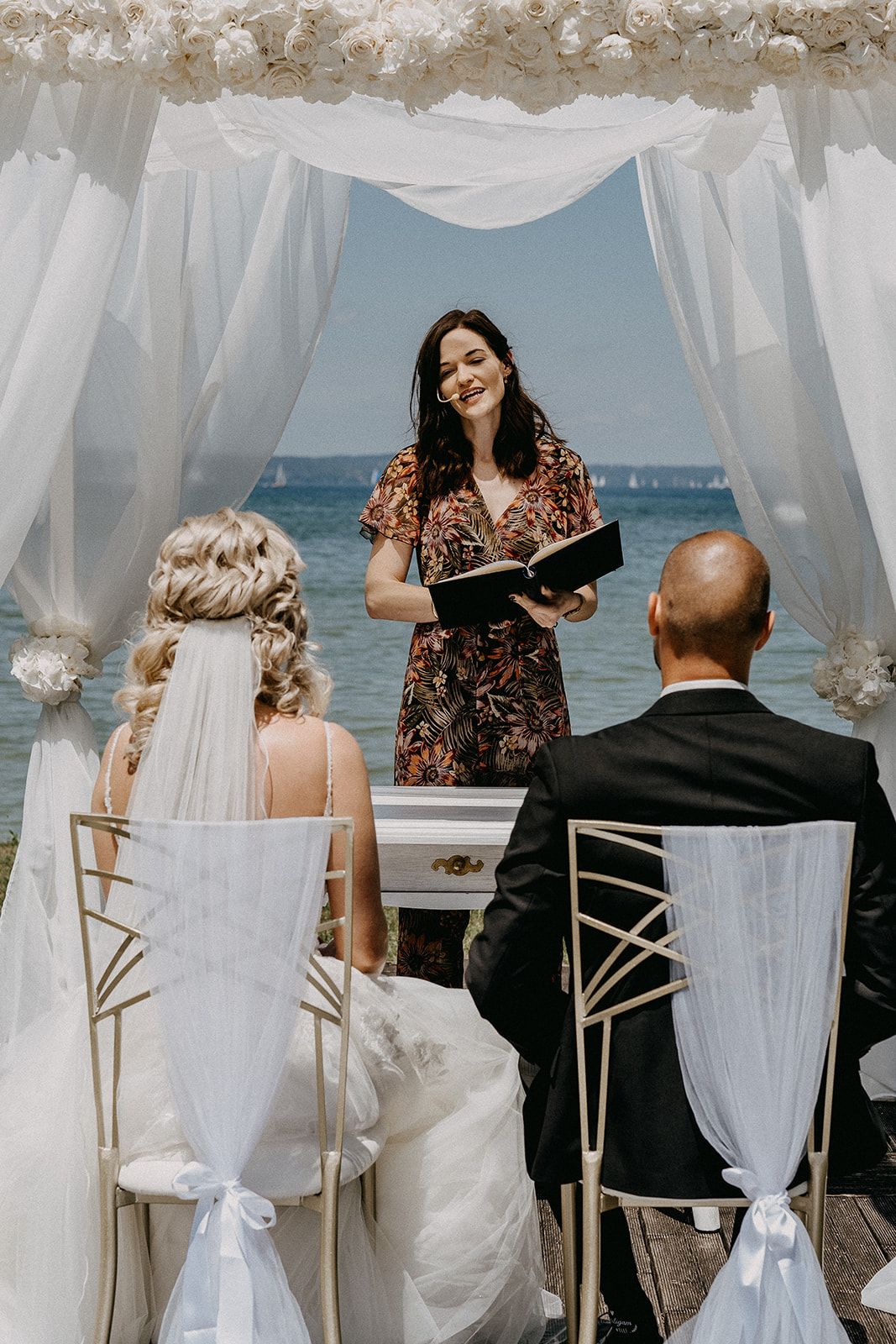 Smiling officiant reads from book as bride and groom sit under floral arch at waterfront ceremony.