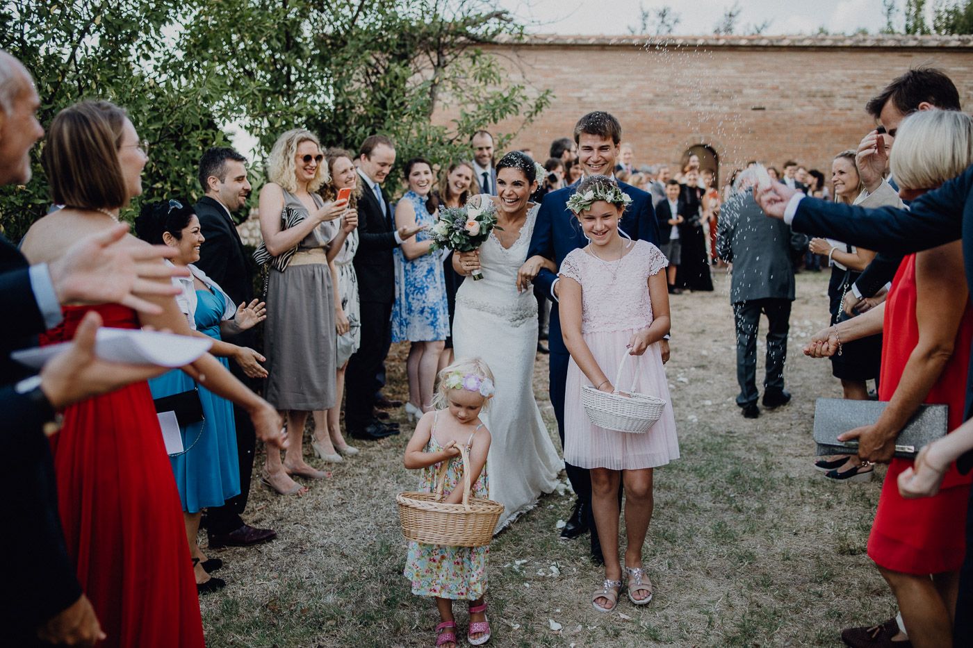 Bride and groom walk through cheering guests with flower girl during festive outdoor wedding reception exit. Tuscany Villa Catignano
