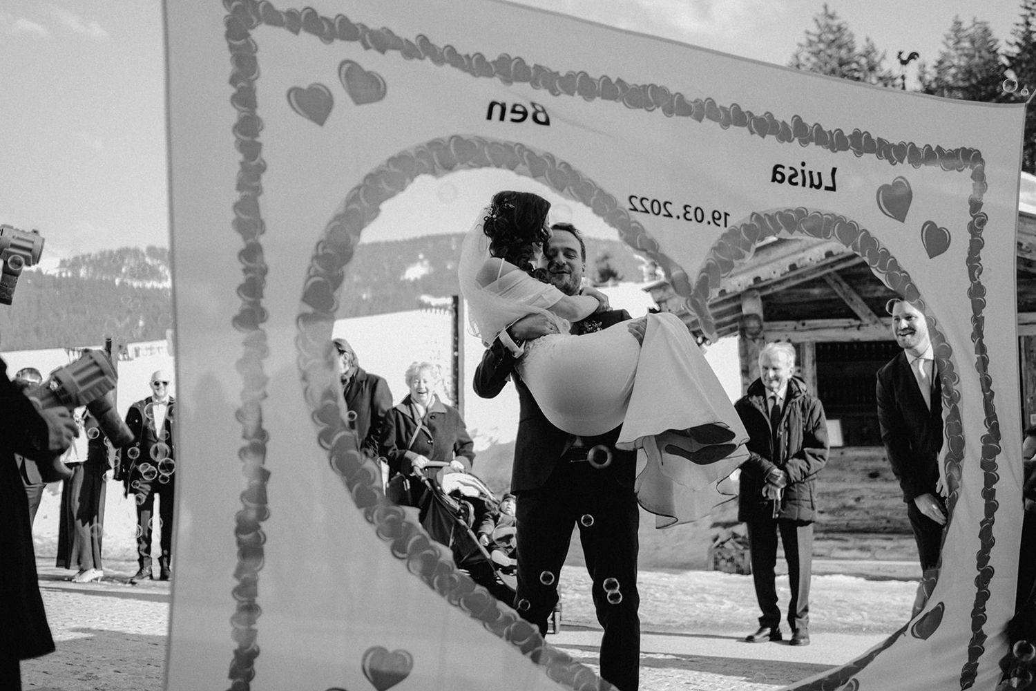 Black and white photo of groom lifting bride through heart-shaped decorative frame at winter mountain wedding celebration. Jufenalm