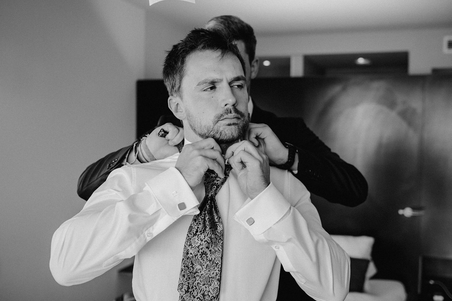 Black and white photo of groom adjusting paisley tie while getting dressed for wedding ceremony.
