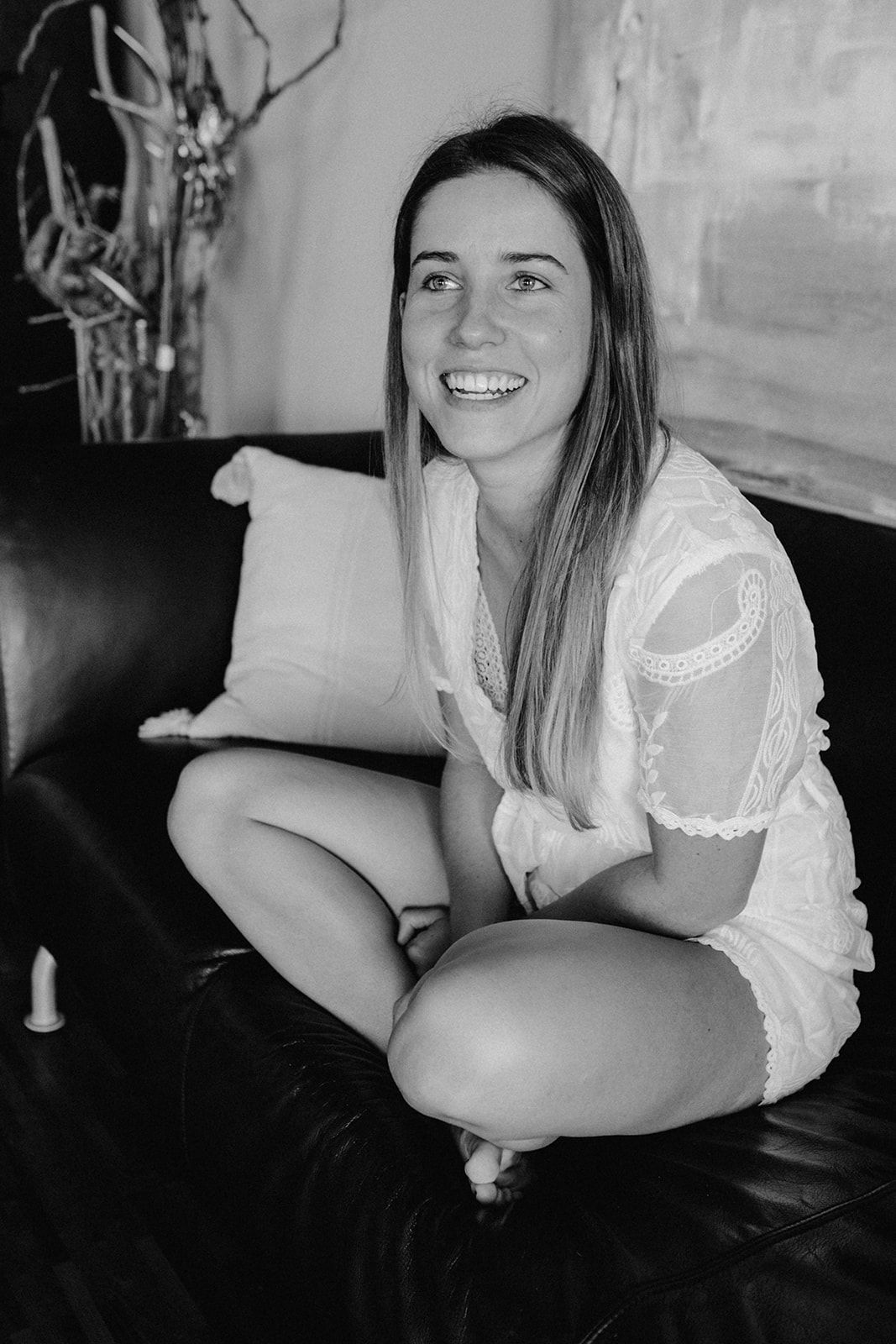 Smiling bride in white lace dress sitting cross-legged on leather sofa in black and white photo.