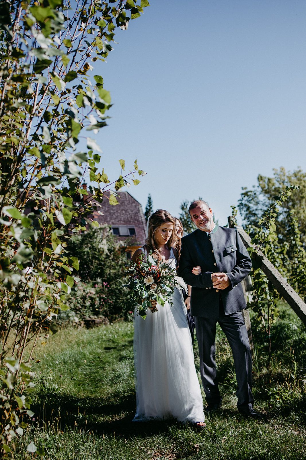 Smiling bride in white dress holds bouquet while walking with older man through green garden.