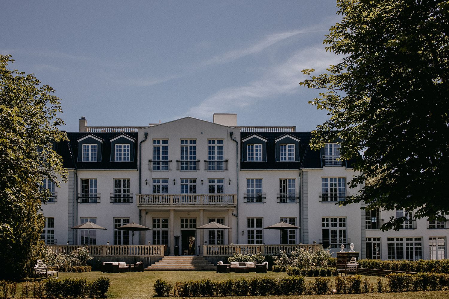 Grand white manor house wedding venue surrounded by lush green trees under blue sky. Hideaway Starnberger See