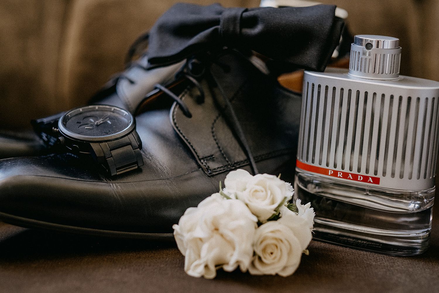 Black leather groom shoes with a watch, white roses boutonniere, and Prada cologne arranged on a surface.