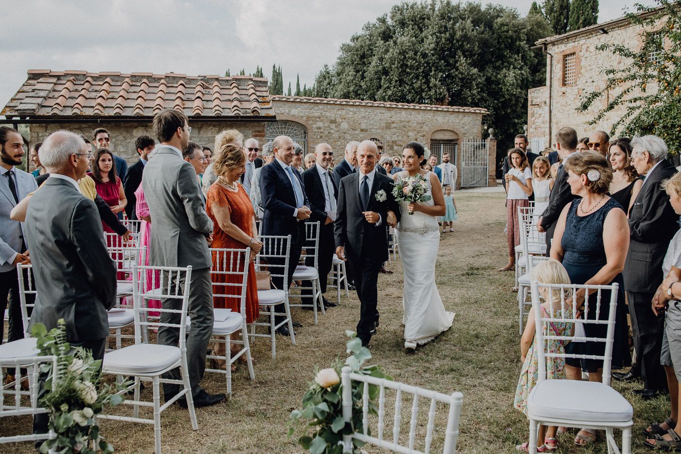 Bride walks down outdoor wedding aisle with escort past seated guests at rustic Tuscan venue. Tuscany Villa Catignano