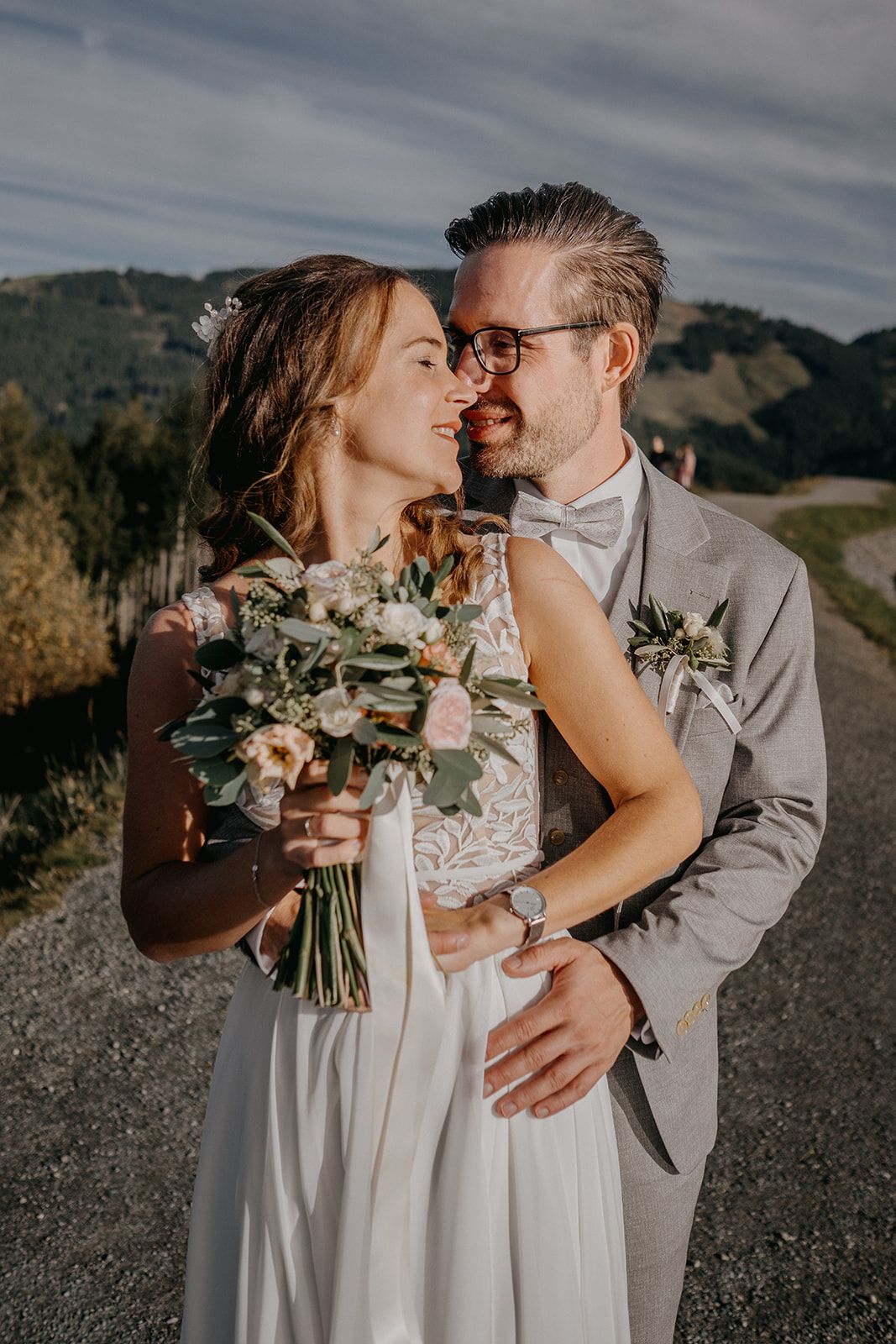 Happy bride and groom share a romantic moment outdoors, bride holding a pastel bouquet with mountain backdrop.
