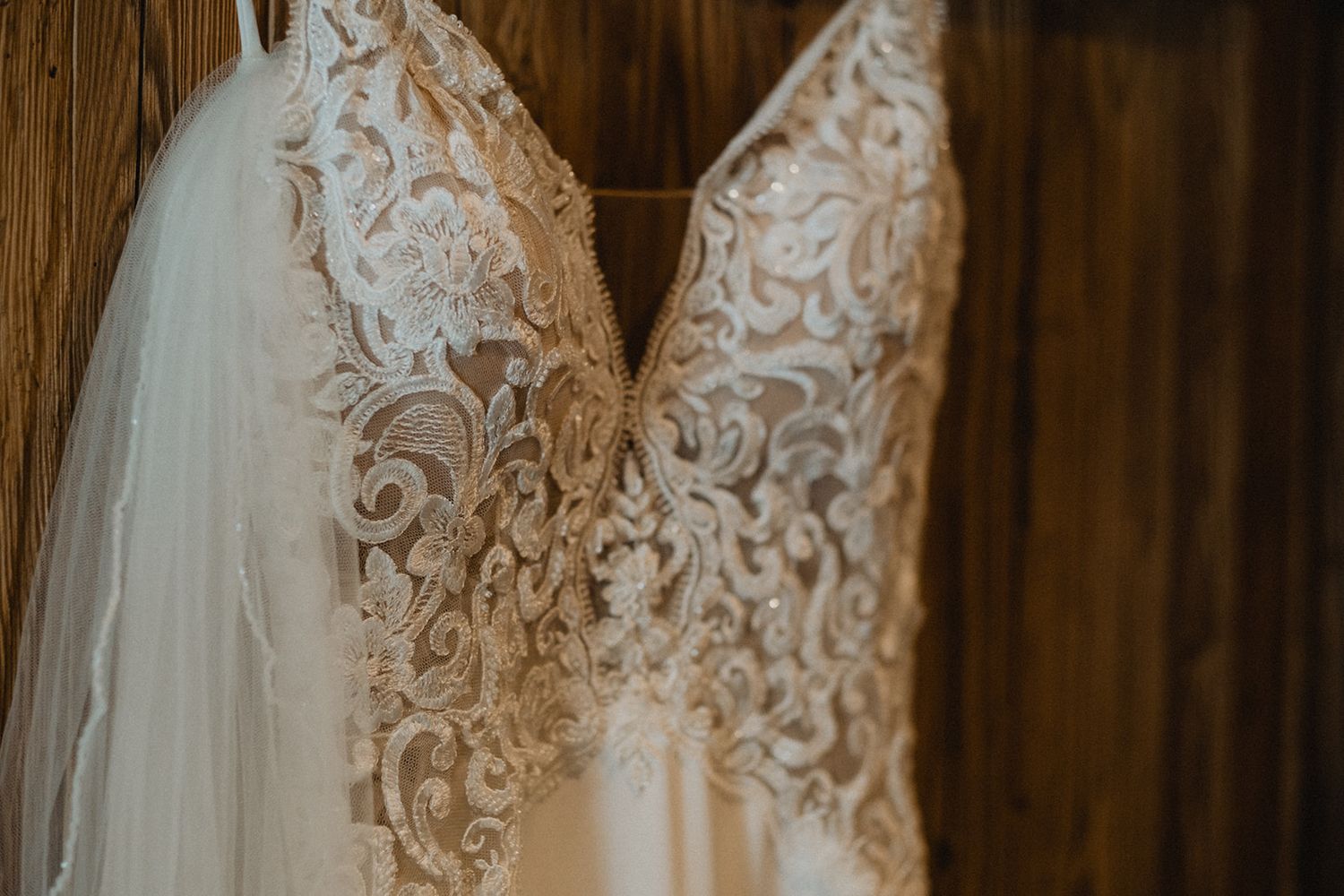Close-up of an elegant lace wedding dress with intricate floral embroidery hanging on a wooden wall.