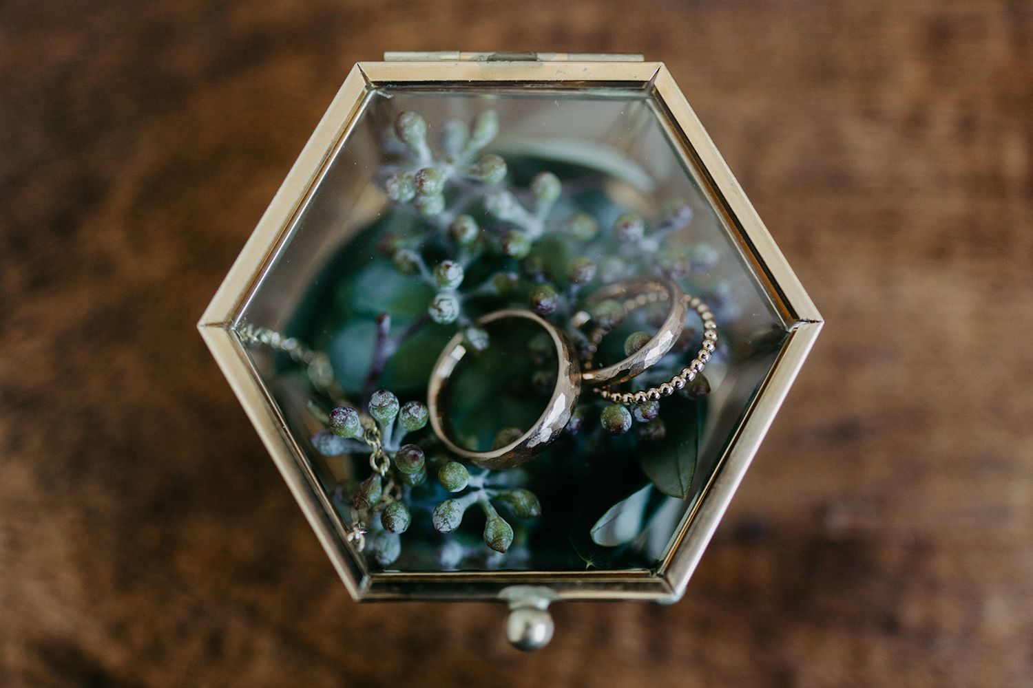 Gold wedding bands and diamond ring displayed in hexagonal glass terrarium box with greenery.