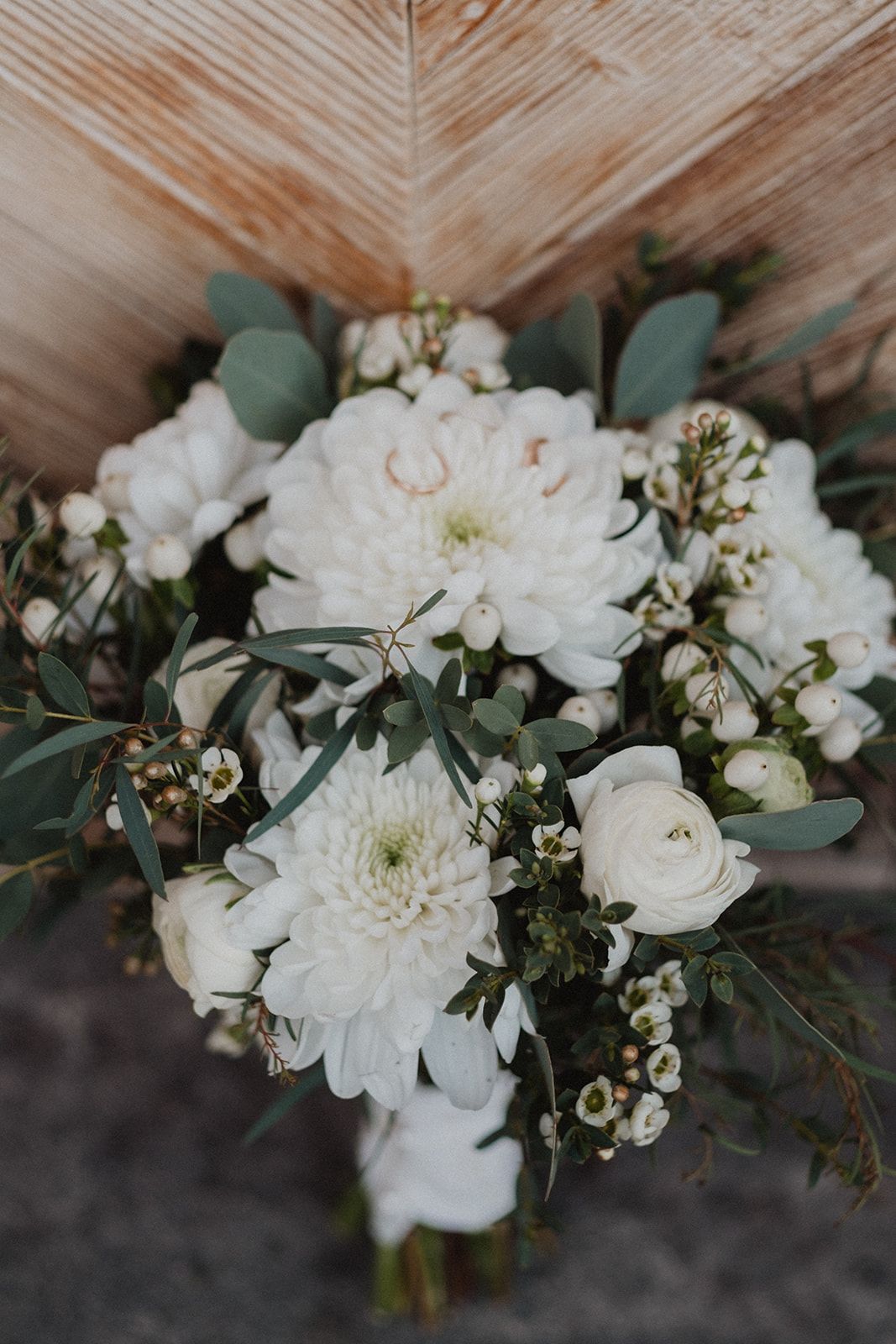 Elegant white wedding bouquet with chrysanthemums, ranunculus, and eucalyptus on rustic wooden backdrop. Jufenalm