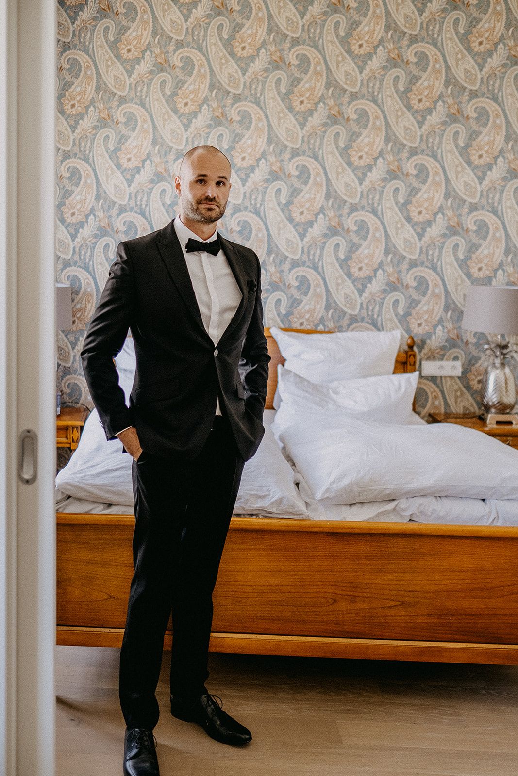 Dapper groom in black tuxedo with bow tie standing confidently in a hotel room with paisley wallpaper.