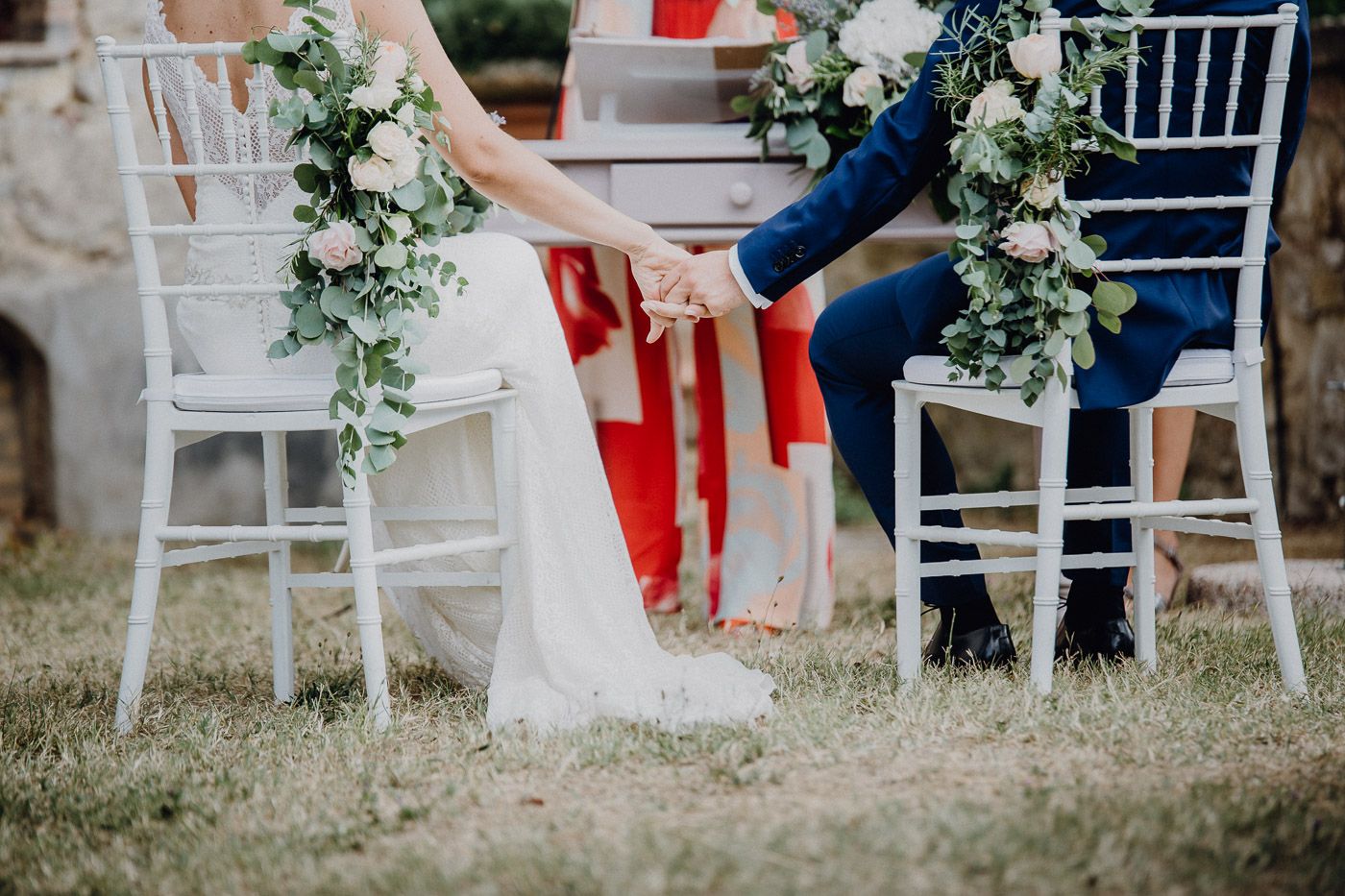 Bride and groom hold hands while seated in white chiavari chairs decorated with greenery at outdoor wedding. Tuscany Villa Catignano