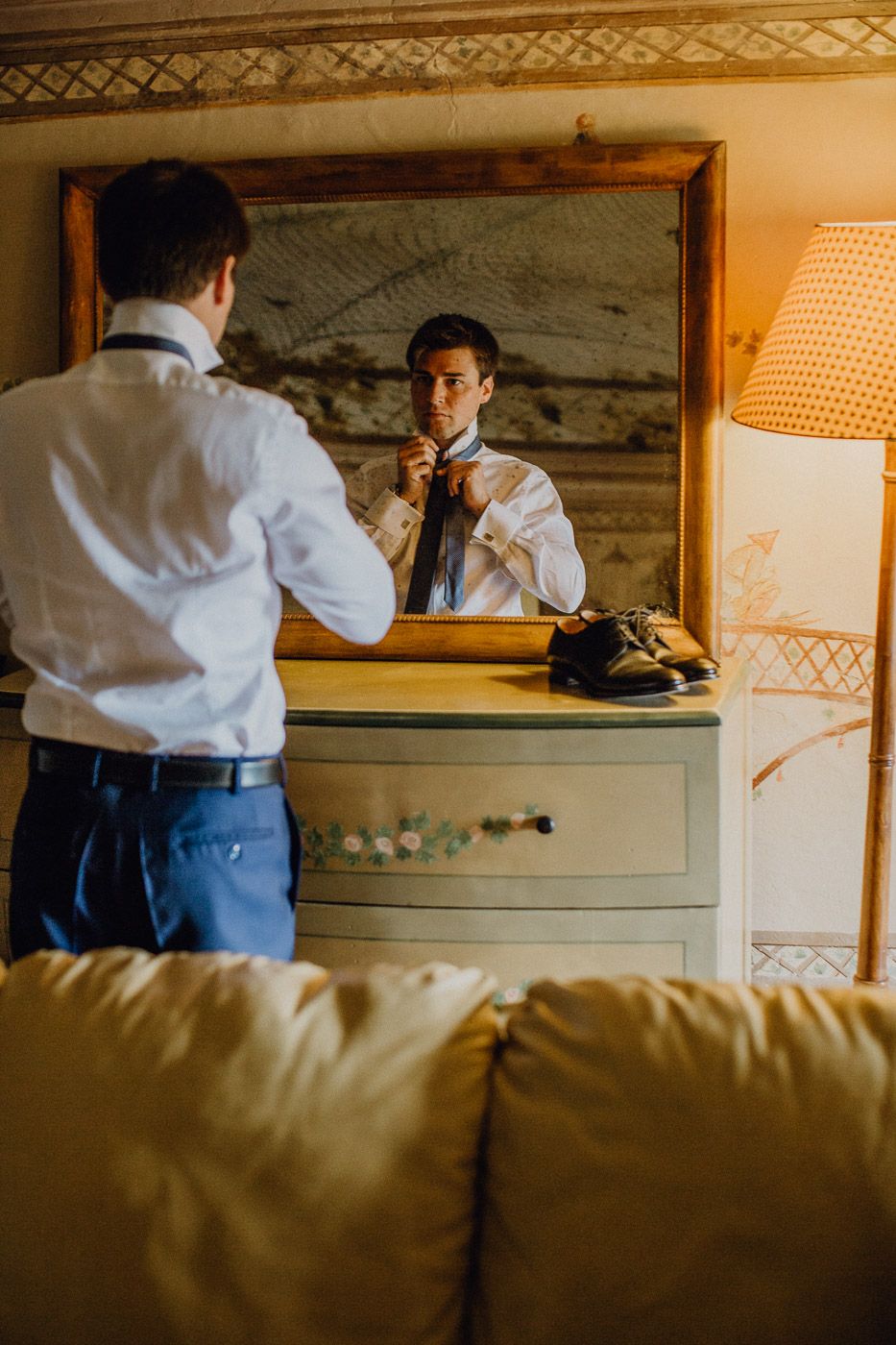 Groom in white shirt adjusts his tie while looking in a mirror during wedding preparations. Tuscany Villa Catignano