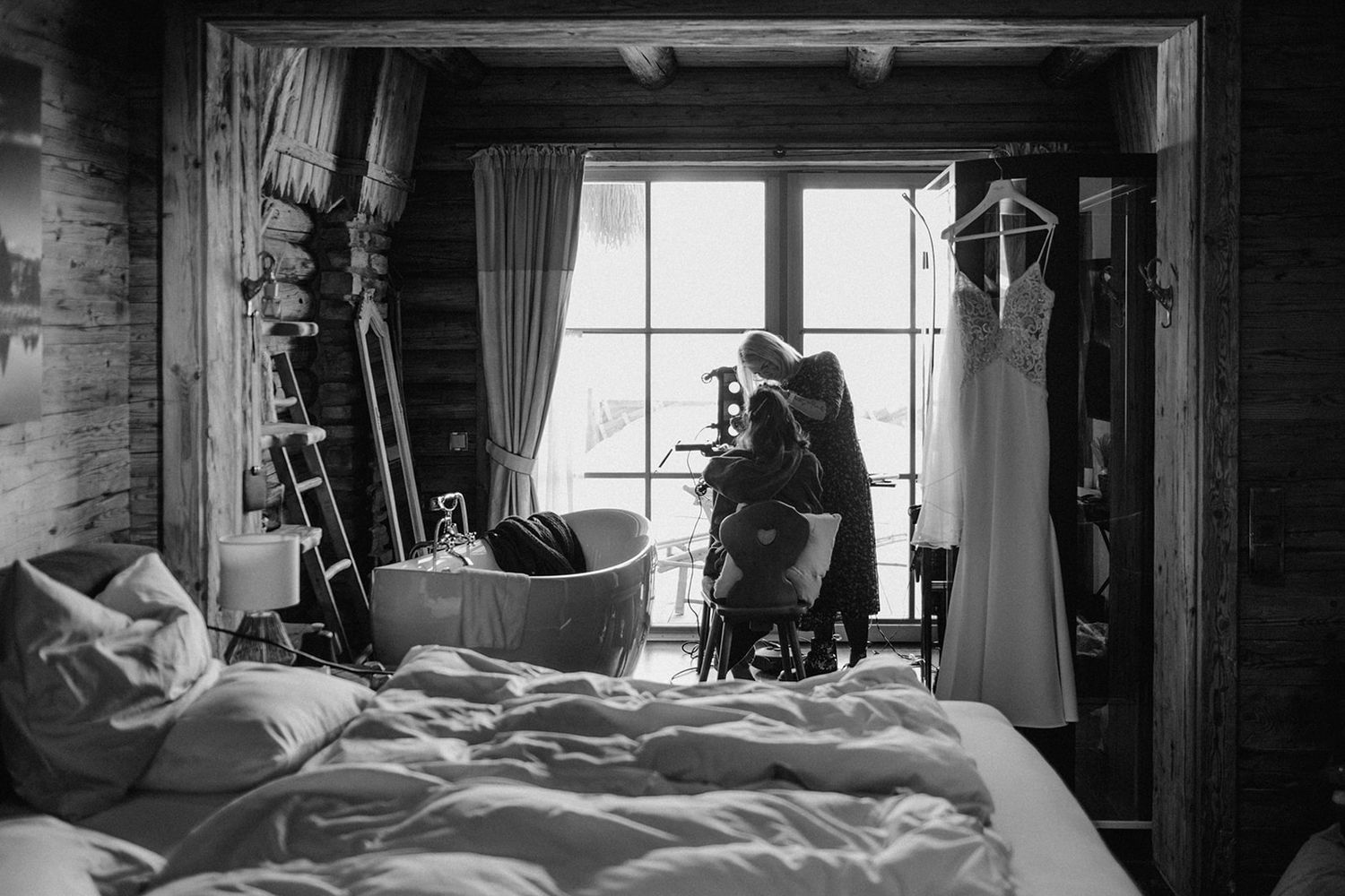 Black and white photo of a bride getting ready near a window with her wedding dress hanging nearby.