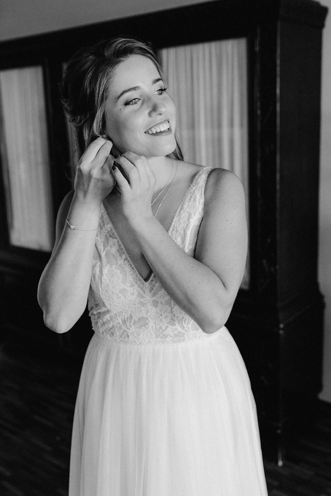 Smiling bride in lace wedding dress putting on earrings, elegant black and white bridal preparation photo.