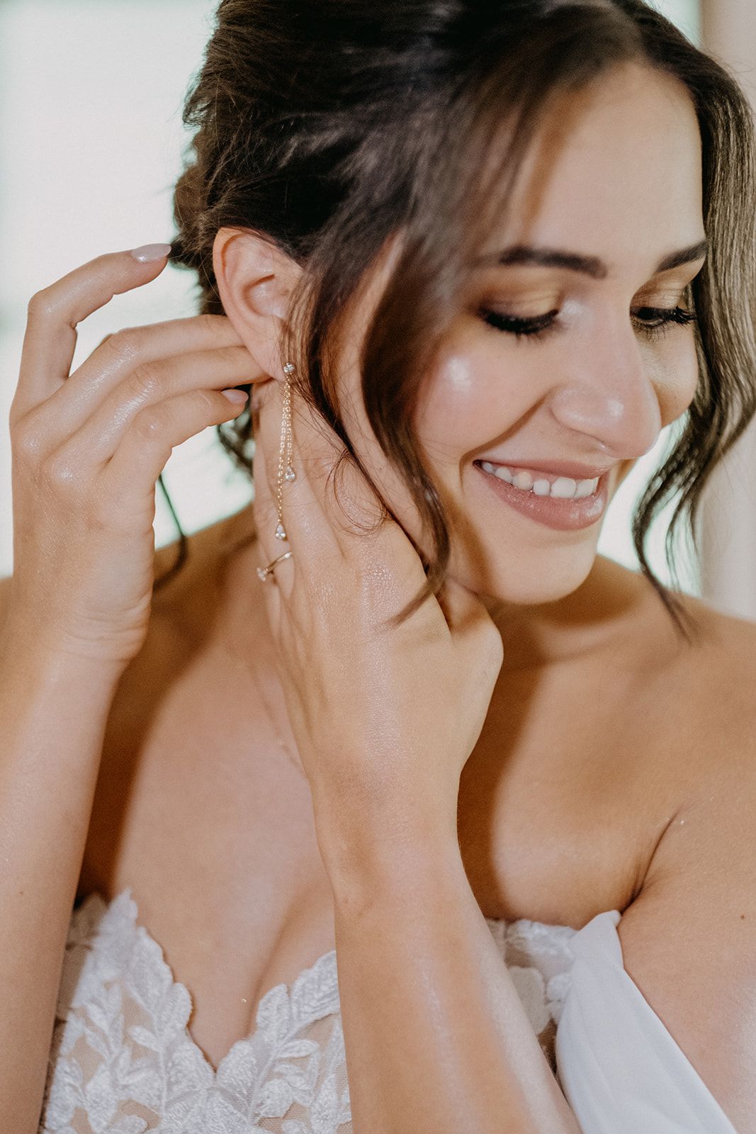 Smiling bride putting on elegant gold drop earrings wearing a lace wedding dress.