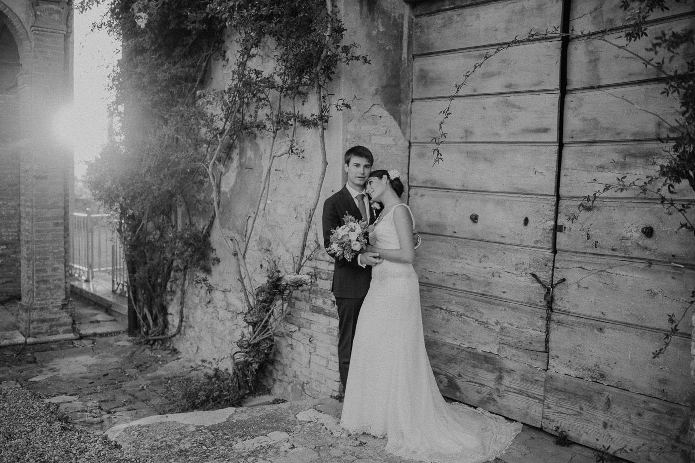 Black and white photo of bride and groom posing beside rustic stone wall with ivy. Tuscany Villa Catignano