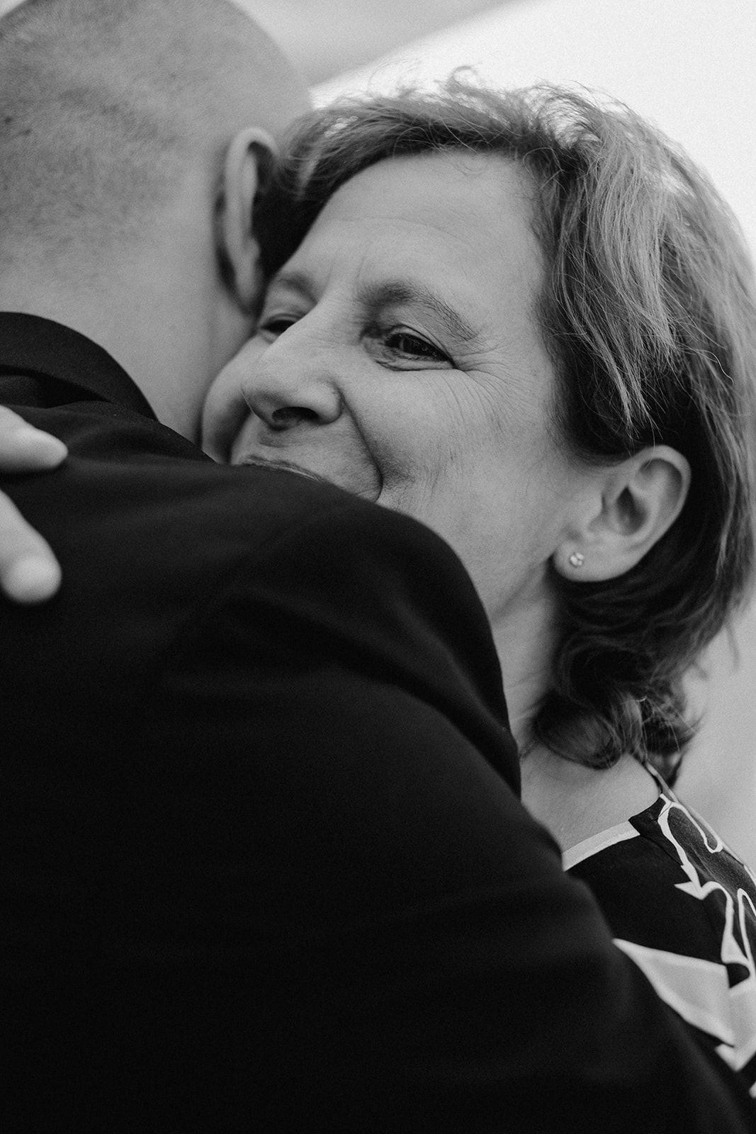 Black and white photo of a woman smiling while sharing an emotional hug at a wedding.