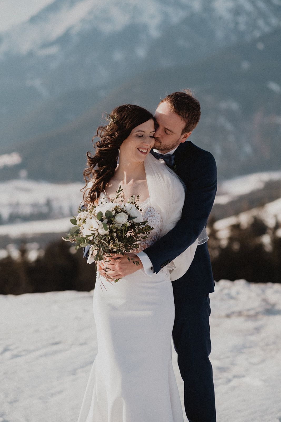 Groom in navy suit embraces bride in white lace dress holding white bouquet against stunning snowy mountain backdrop. Jufenalm