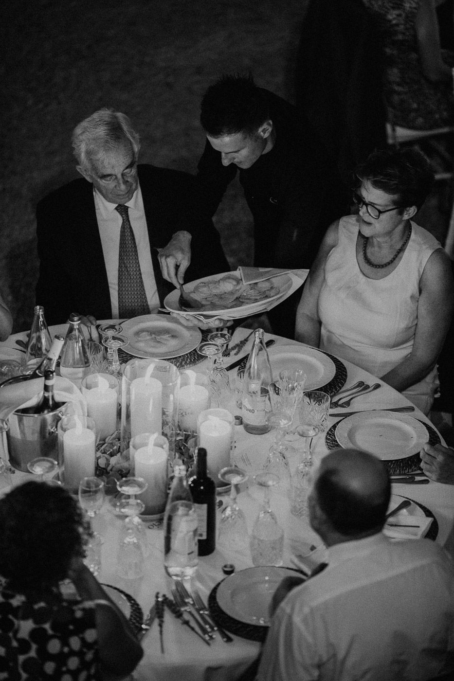 Black and white photo of wedding guests being served food at candlelit reception dinner table. Tuscany Villa Catignano
