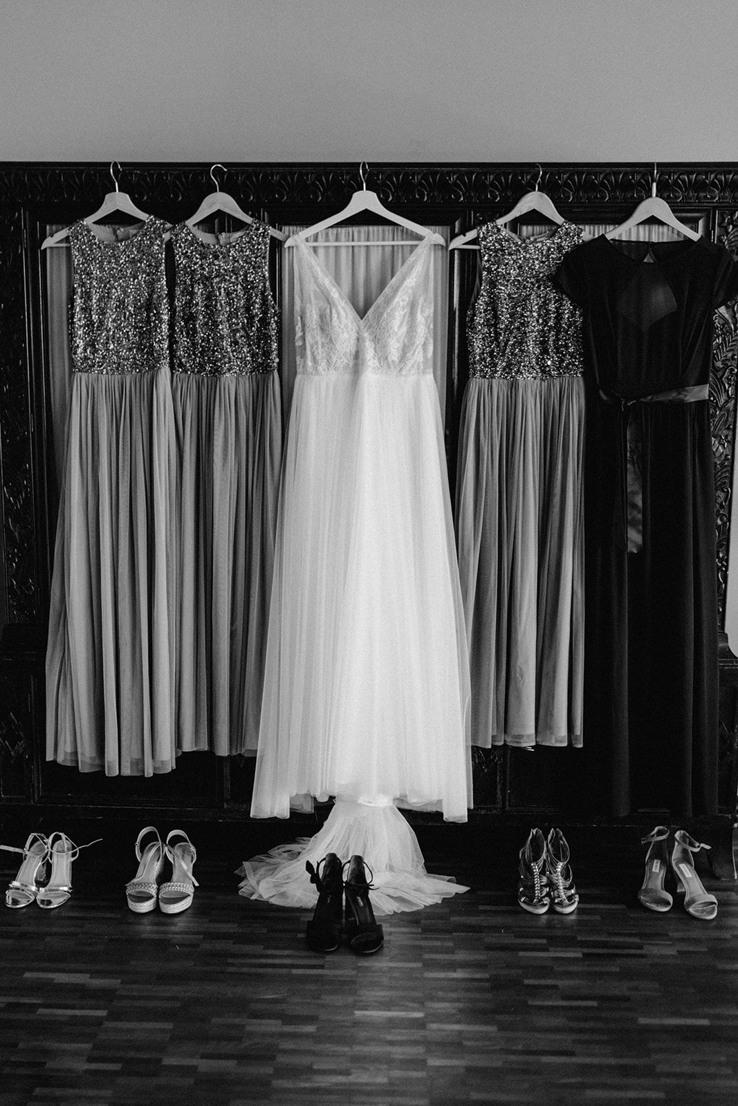 Black and white photo of wedding dress and sequined bridesmaid gowns hanging with heels below.