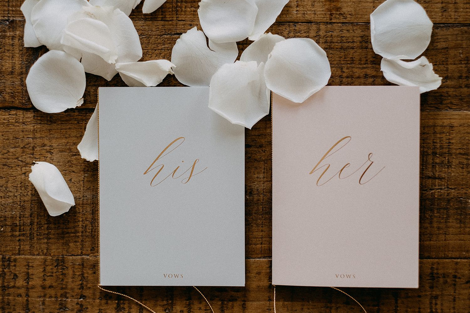 His and her wedding vow books in gray and blush pink with gold script, surrounded by white rose petals.