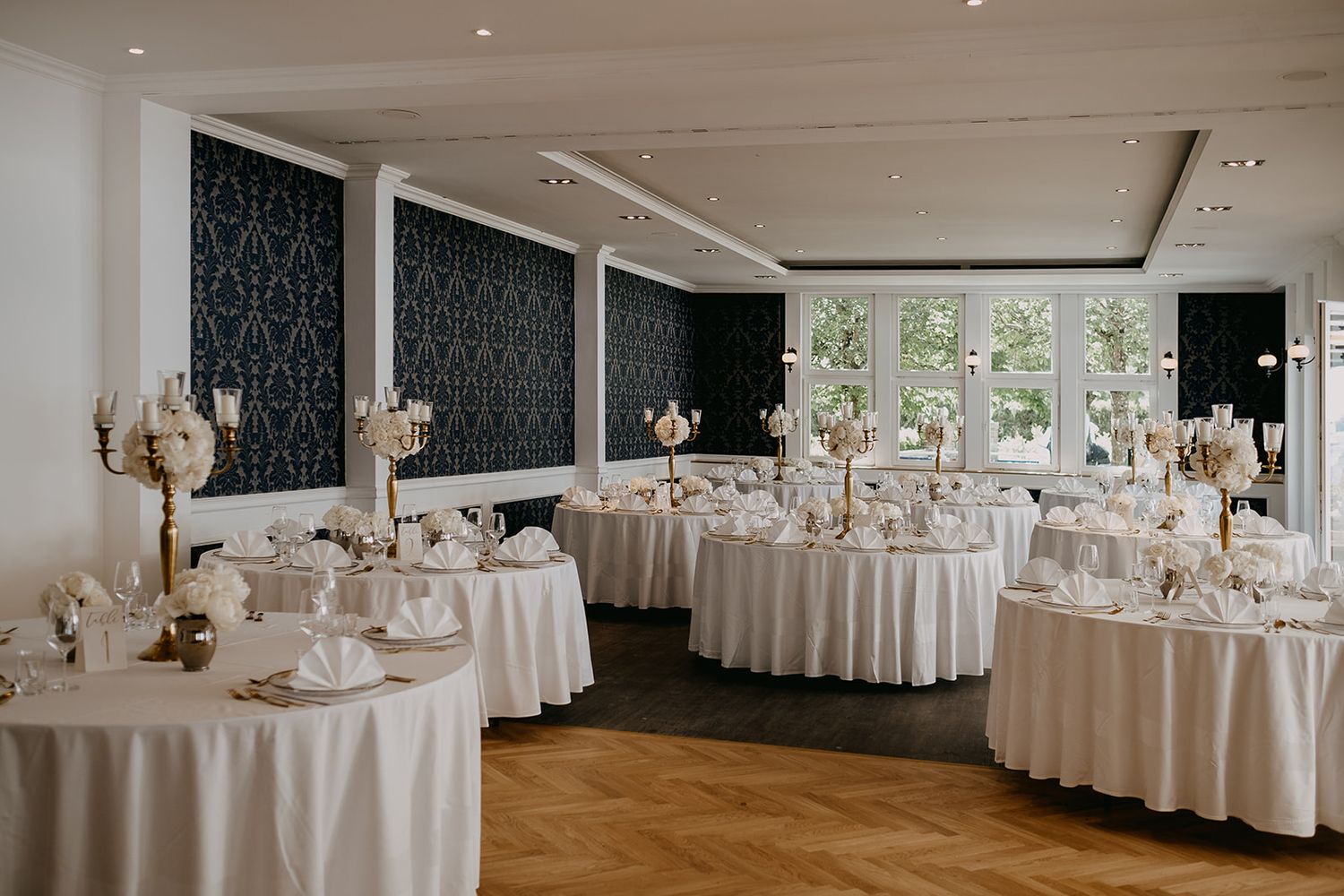 Elegant wedding reception hall with round white-draped tables, gold candelabras, and decorative dark patterned walls.