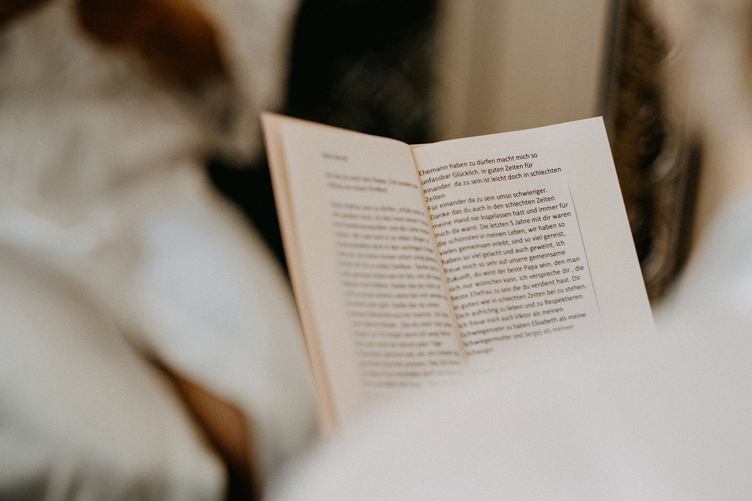 Bride in white dress reading a book with soft bokeh background on wedding morning.