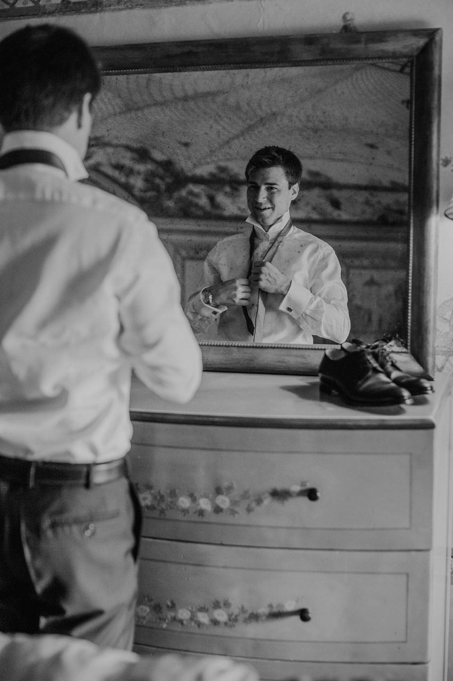 Black and white photo of smiling groom adjusting his tie in a mirror before the wedding. Tuscany Villa Catignano