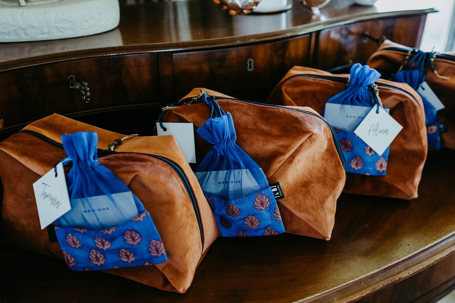 Brown suede groomsmen gift bags with blue floral pouches and personalized tags on wooden table.