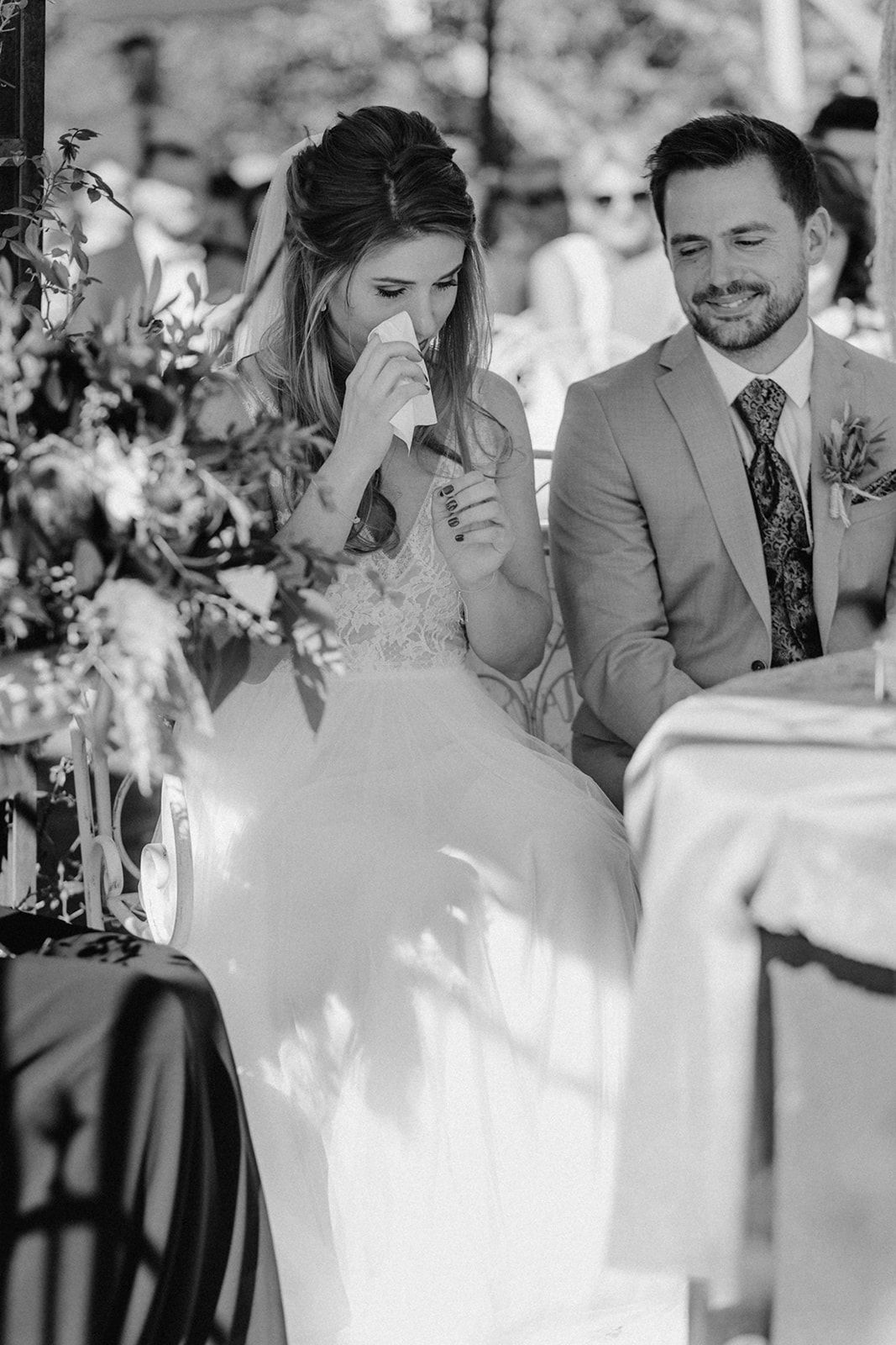 Black and white photo of emotional bride dabbing tears while groom smiles at outdoor wedding.