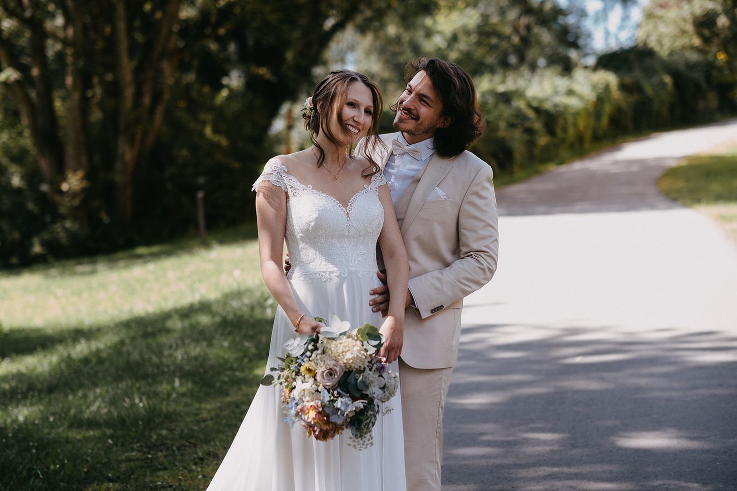 Smiling bride and groom walking on sunny path, bride holding wildflower bouquet.