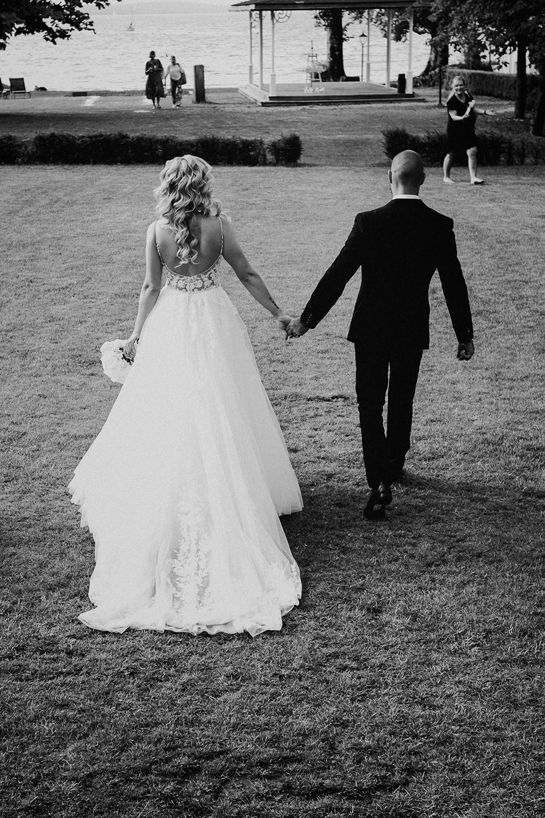 Black and white photo of bride and groom holding hands walking across a lawn toward a waterfront.