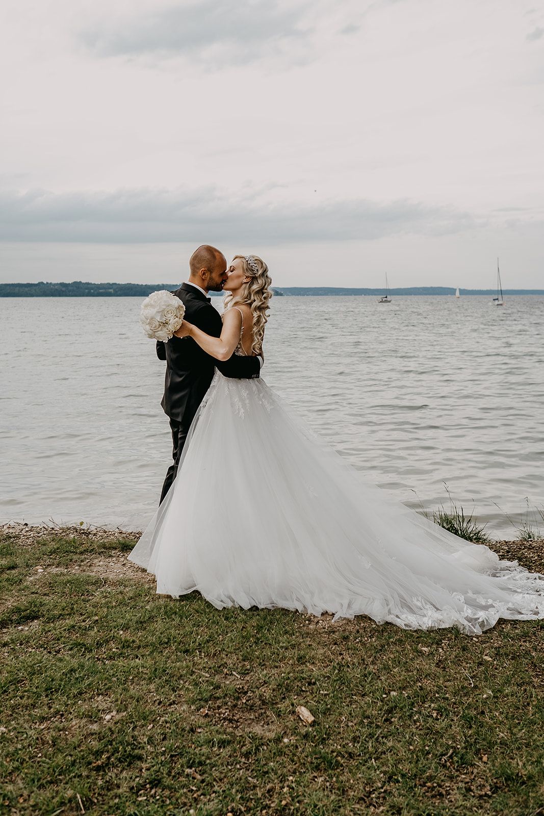 Bride and groom share a romantic kiss by the lakeside under a cloudy sky on their wedding day.