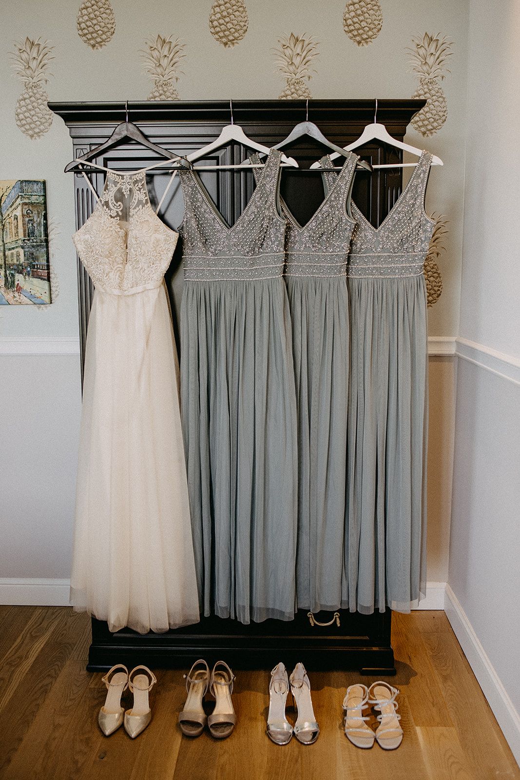 White wedding gown and three grey bridesmaid dresses hang on a rack above matching bridal heels.