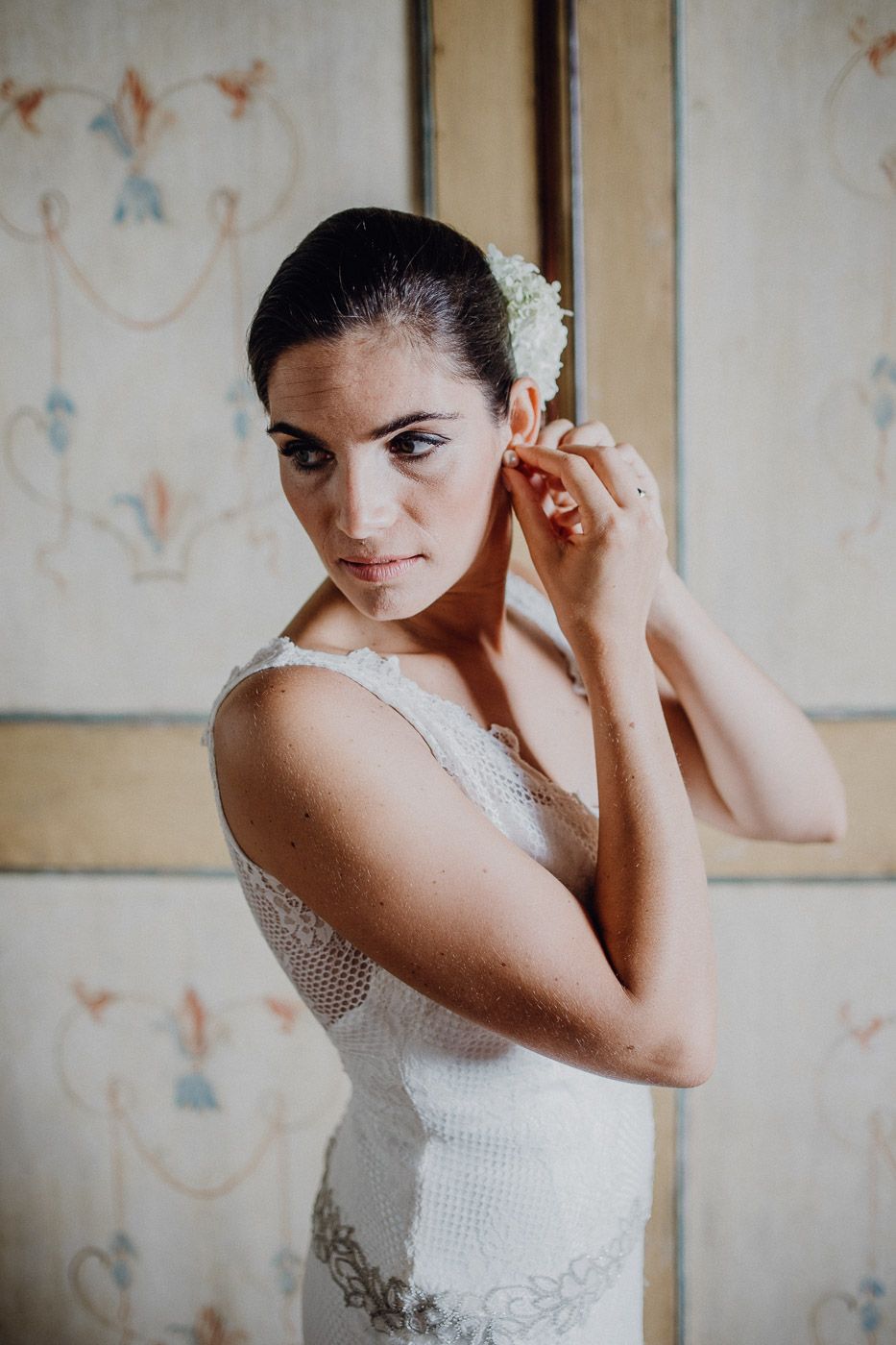 Elegant bride in lace wedding gown places a white flower accessory in her hair. Tuscany Villa Catignano