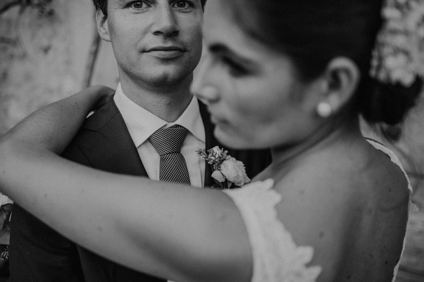 Black and white close-up of groom looking at camera while bride embraces him outdoors. Tuscany Villa Catignano