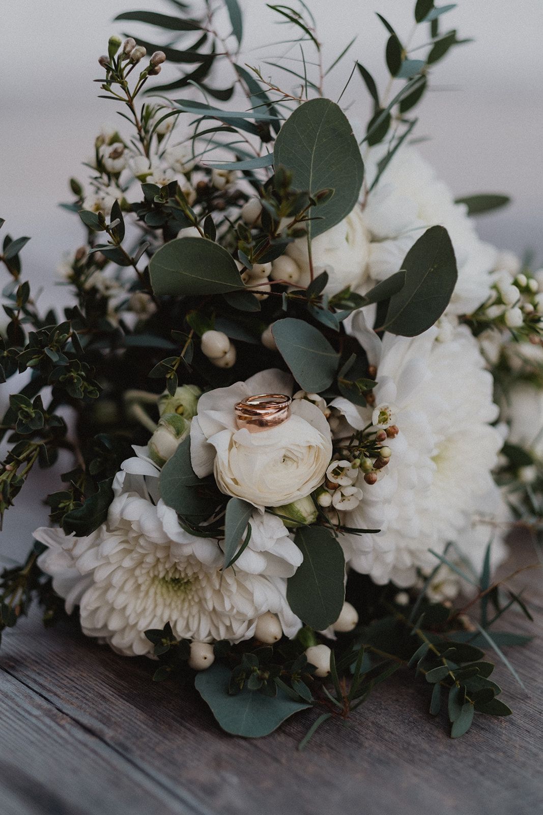 Elegant bridal bouquet of white ranunculus, chrysanthemums, and eucalyptus with two gold wedding rings.