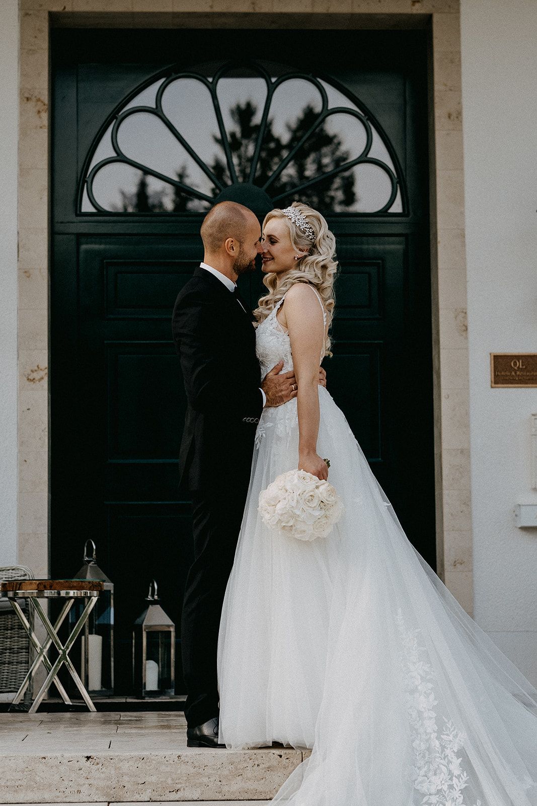 Bride and groom share a tender forehead touch before a dark ornate door, holding white bouquet.