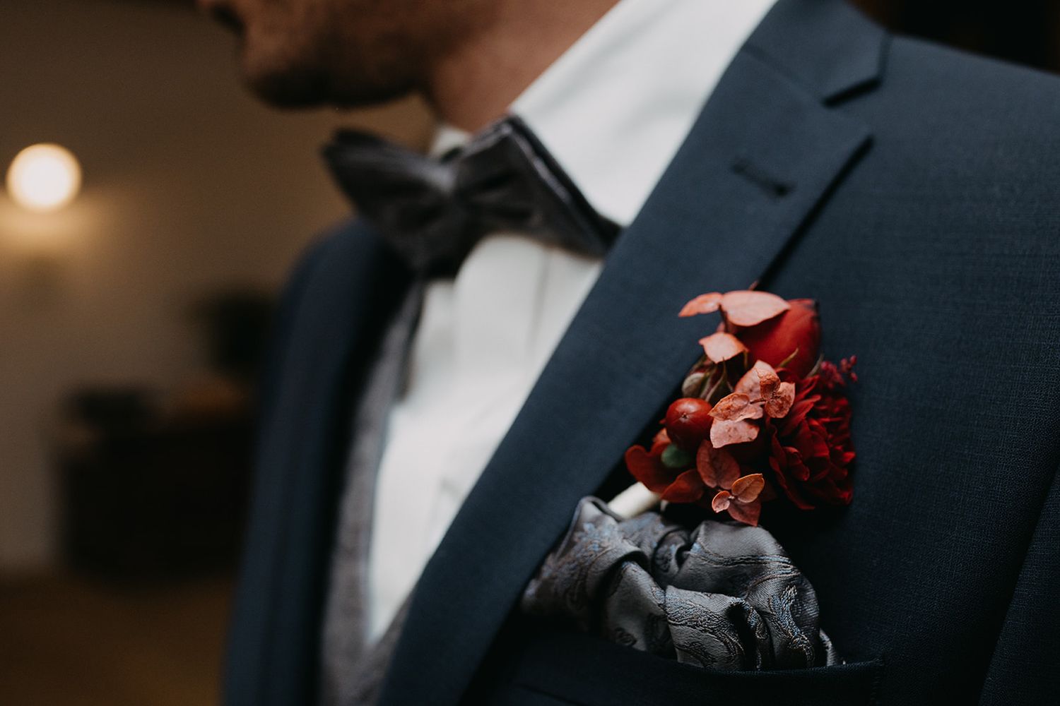 Groom in navy suit with red floral boutonniere and black bow tie at wedding.