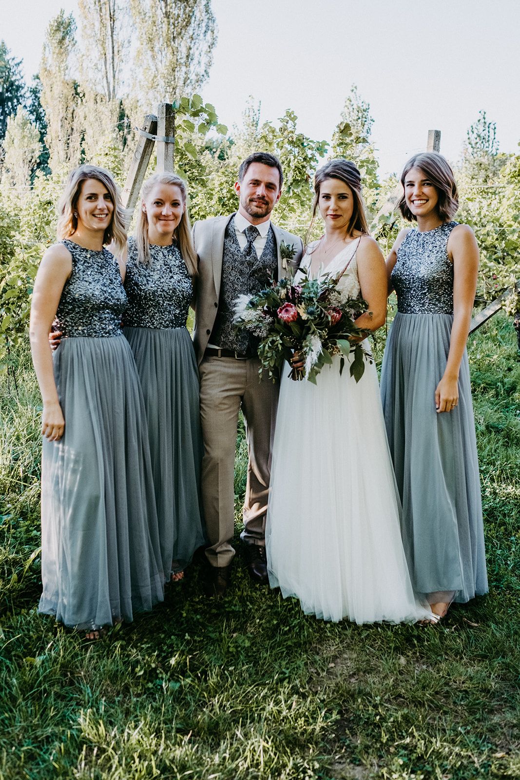 Wedding party posing outdoors with bride in white gown and three bridesmaids in sequined steel-blue dresses.